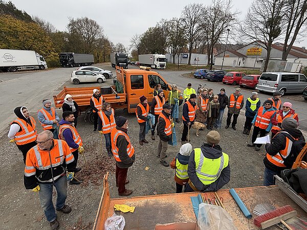 Eine Gruppe von Menschen mit Warnwesten steht in einem Halbkreis um einen kleinen organeben LKW herum. Es sind ehrenamtlich die bei einer Müllsammelaktion der Stadt Alfeld mit machen wollen. Die Menschen sind jeden Alters. 